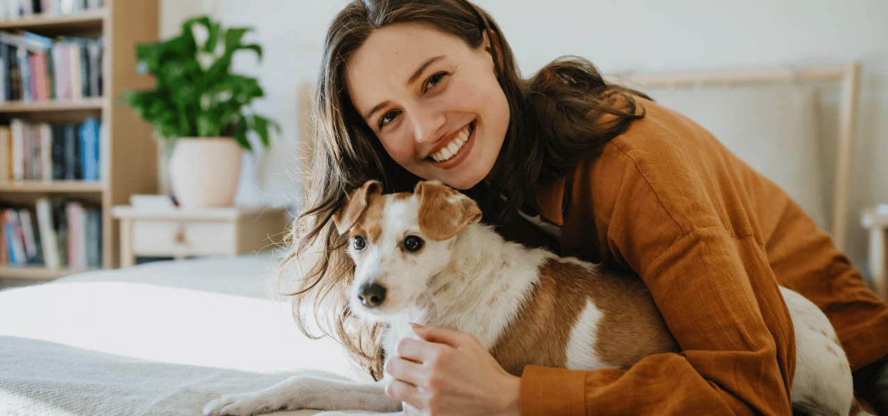 Woman holding a small dog in a cozy room with books and plants.